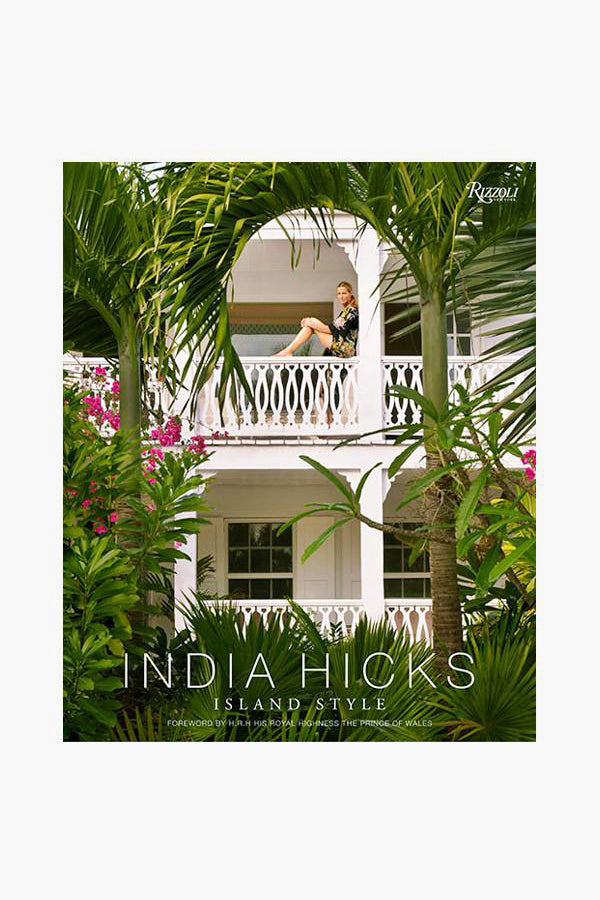 A woman relaxes on a balcony of a white two-story house with palm trees and pink flowers. The text reads, Rizzoli presents "India Hicks Island Style," showcasing relaxed island living and bohemian décor from Rizzoli.