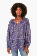 A woman wears a loose, long-sleeve purple floral Iris Wildflower Bardot Top with ruffled collar from Love Tomas by Emerson Fry, paired with light blue jeans. The top is crafted from soft organic cotton.