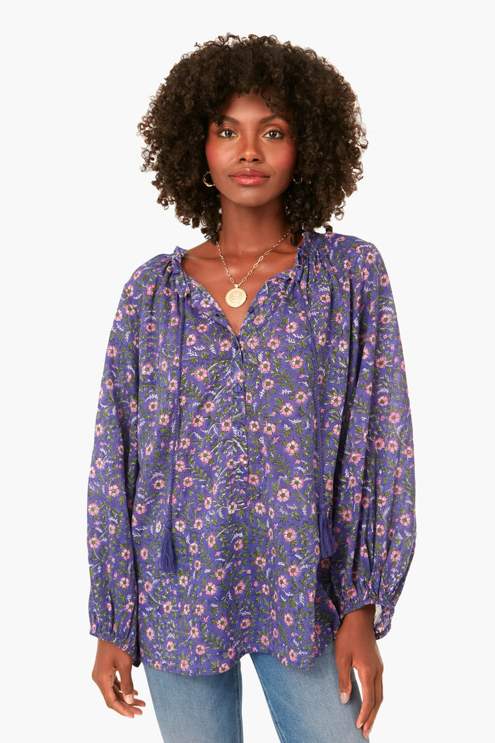 A woman wears a loose, long-sleeve purple floral Iris Wildflower Bardot Top with ruffled collar from Love Tomas by Emerson Fry, paired with light blue jeans. The top is crafted from soft organic cotton.