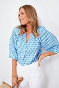 A woman wearing a Vineyard Vines blue and white patterned gauzy cotton top with puff sleeves and a ruffle collar, paired with white pants and holding a straw hat.