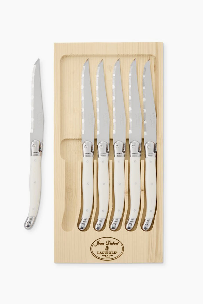 A Laguiole wooden box set with six handmade steak knives featuring white handles, crafted in France, plus one knife positioned separately on the left side of the box.