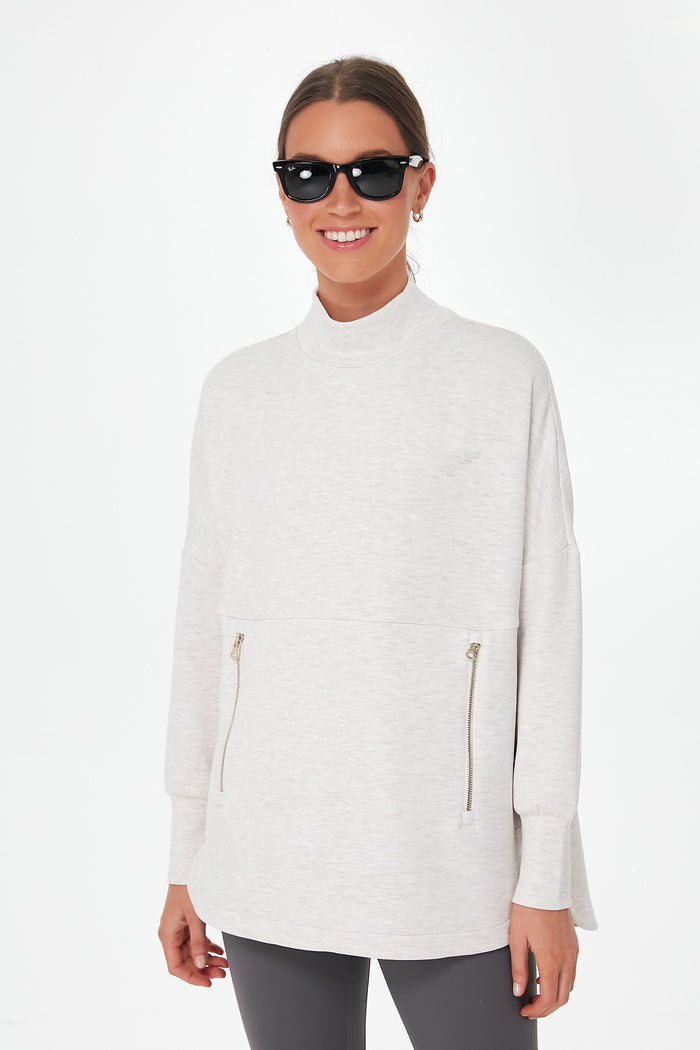 A woman in Varley activewear—a light mock neck zip sweatshirt and dark leggings—smiles at the camera, wearing dark sunglasses.