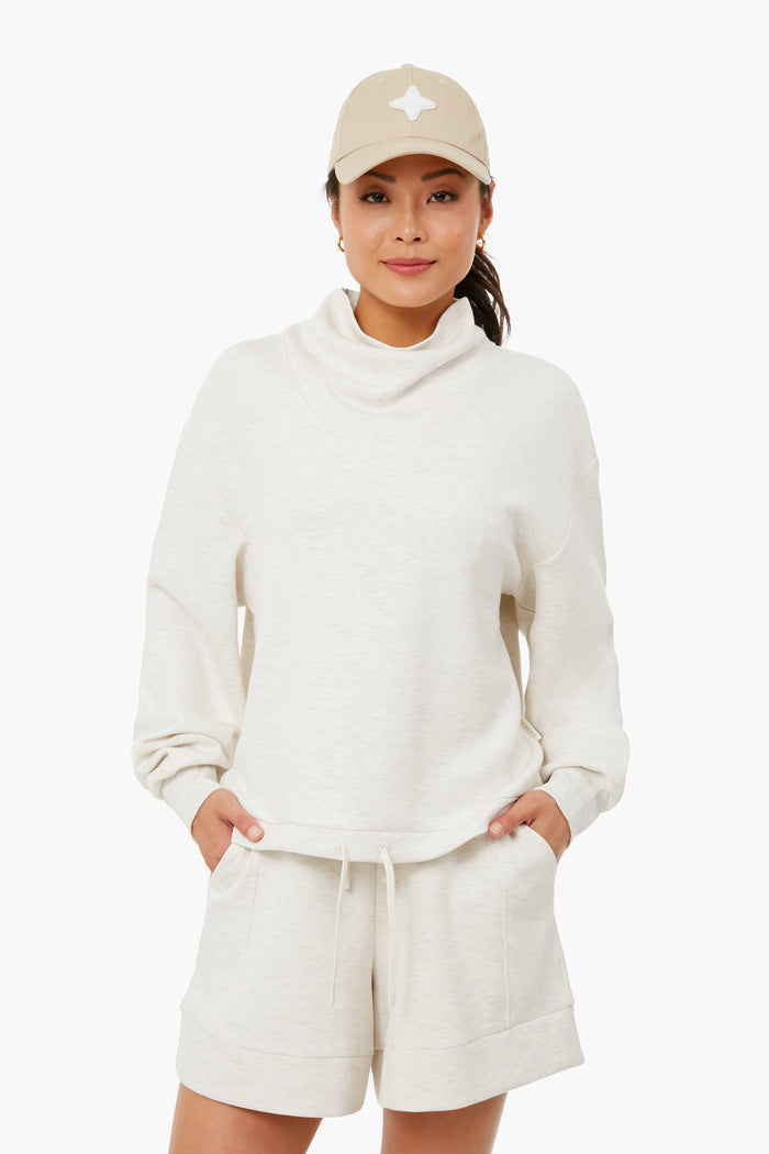 A woman wears Varley’s light beige cowl neck sweatshirt and matching shorts, paired with a beige cap featuring a white shape, her hands in her pockets. The outfit’s lightweight fabric ensures a relaxed, comfortable look.