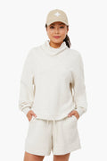 A woman wears Varley’s light beige cowl neck sweatshirt and matching shorts, paired with a beige cap featuring a white shape, her hands in her pockets. The outfit’s lightweight fabric ensures a relaxed, comfortable look.