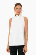 A woman wears a sleeveless white Tuckernuck blouse with a ruffle-neck and stand collar, black pants, gold hoop earrings, and a gold bracelet.