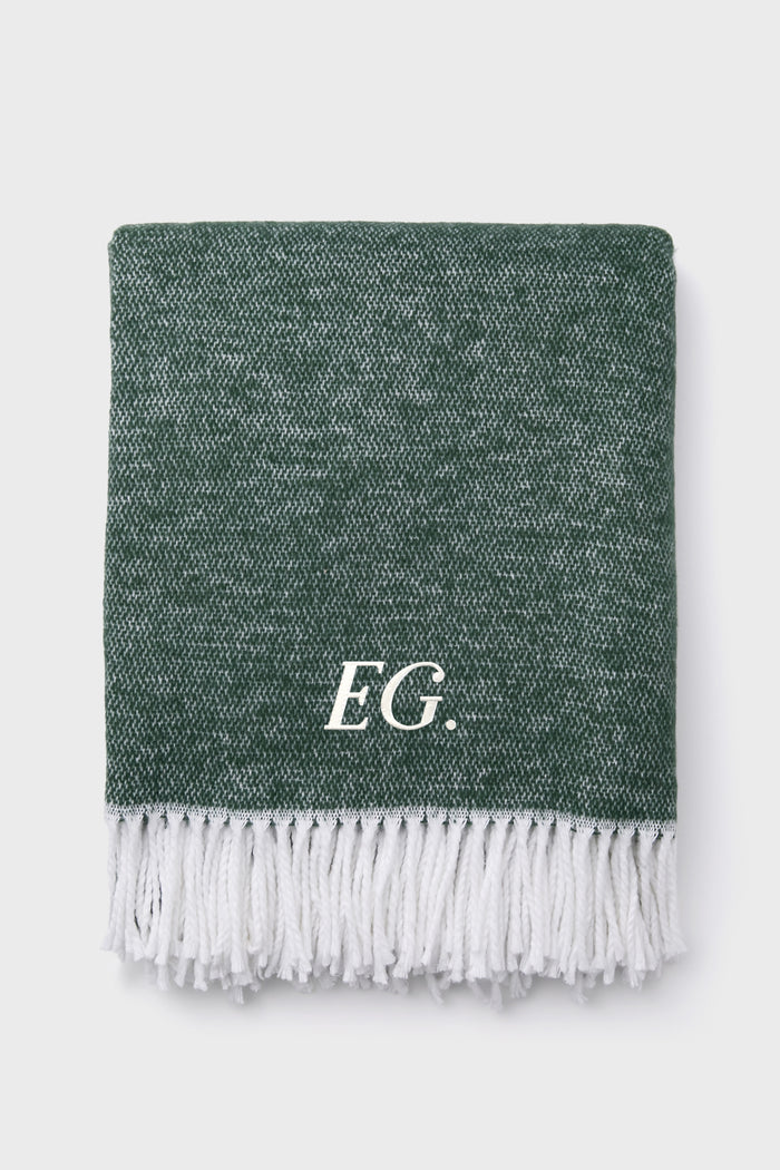 A folded green Jade Pezzo Throw by Matouk, featuring a textured finish, white fringed edges, and the initials E.G. in white on one side, adds an elegant touch.