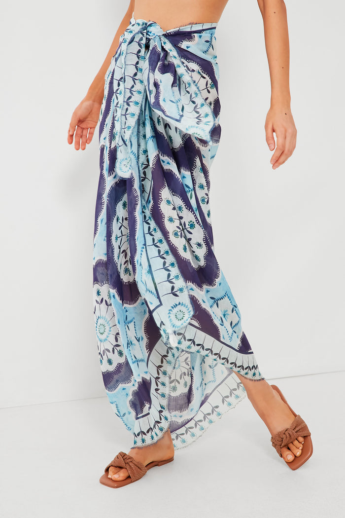 A person wears the ASHA x Tuckernuck blue patterned wrap skirt tied at the waist with brown slip-on sandals, standing confidently and highlighting stylish comfort.