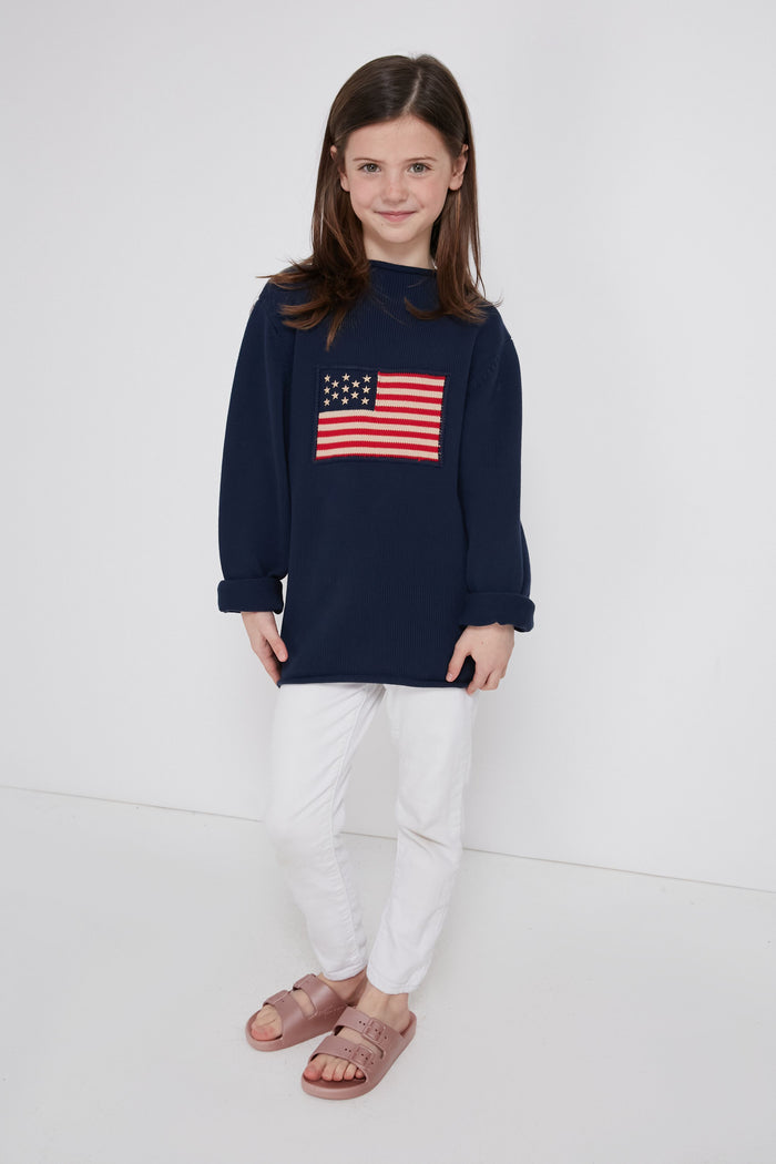 A young girl models a Tuckernuck Tots Kid's Navy Americana Sweater featuring an American flag, styled with white pants and pink sandals—ideal toddler apparel for patriotic occasions.