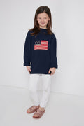 A young girl models a Tuckernuck Tots Kid's Navy Americana Sweater featuring an American flag, styled with white pants and pink sandals—ideal toddler apparel for patriotic occasions.