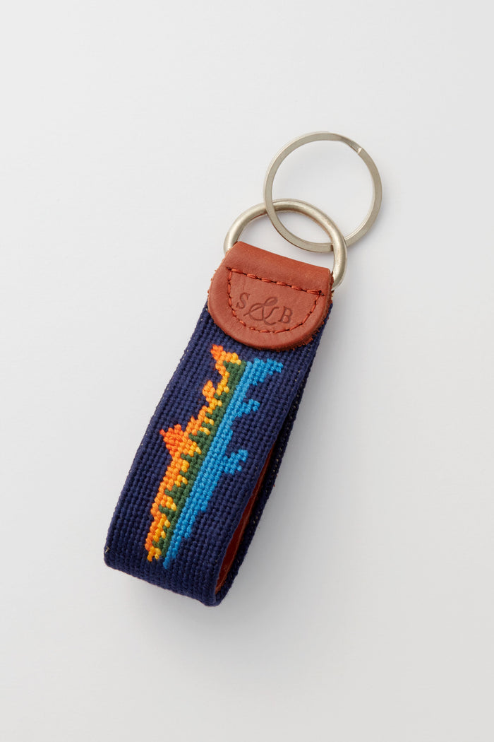 A Smathers and Branson leather key fob with two metal rings, a navy blue fabric band adorned with a colorful embroidered design, and a brown leather accent embossed with initials.