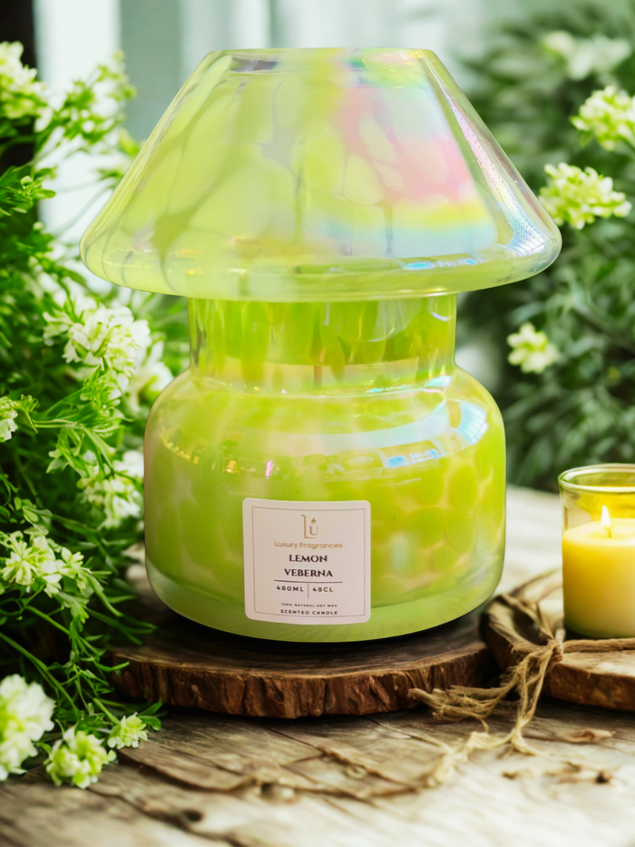 A large jar candle with a translucent iridescent cap and a white label reading 'Lemon Verbena" , sits on a rustic wooden slice. A smaller lit candle is beside it, with green foliage and small white flowers in the background.