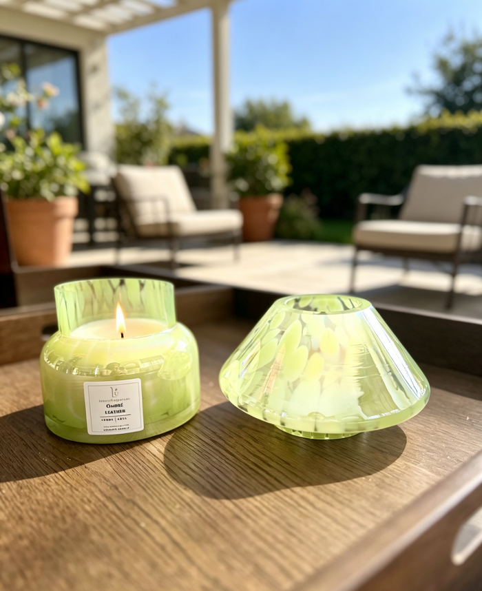 A lit candle in a translucent light green glass jar sits on a wooden table outdoors, with the matching green glass lid placed beside it. The background shows a blurred patio setting with chairs, plants, and a house exterior.