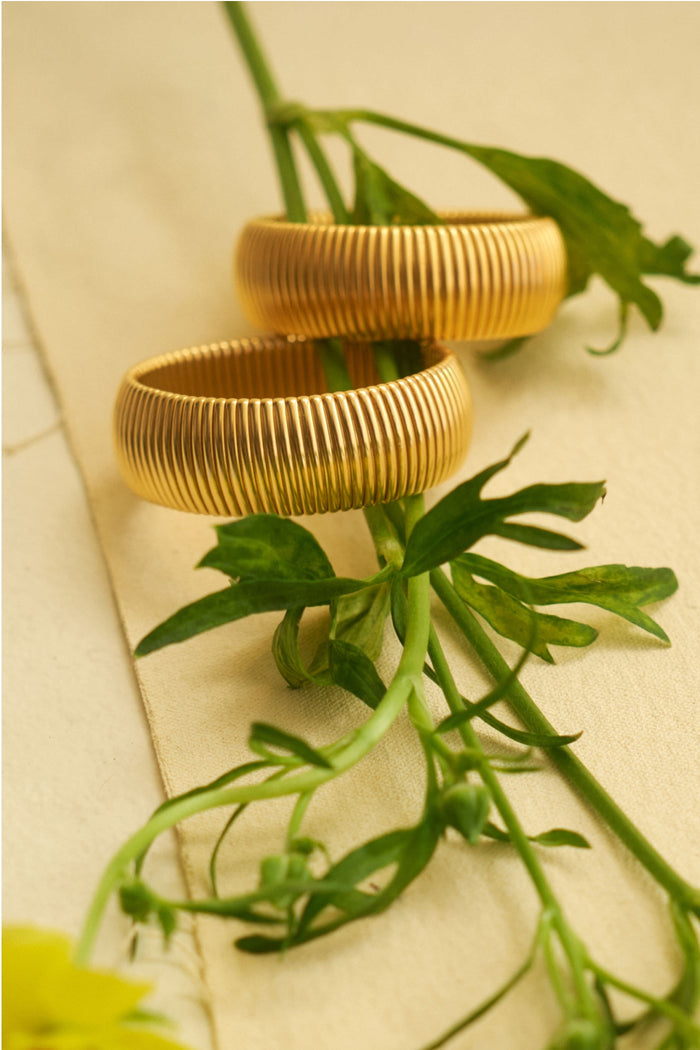 Two textured gold rings rest on green plant stems atop beige fabric, paired with a striking statement bracelet by Ben Amun.