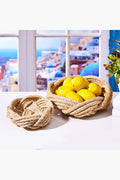 Two hand woven rope baskets by Two's Company sit on a white surface—one filled with yellow lemons, the other empty. A window with a scenic view is nearby.