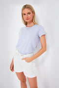 A woman wears a light blue super-soft Marine Layer t-shirt, white high-waisted shorts, and gold jewelry, with one hand in her pocket.