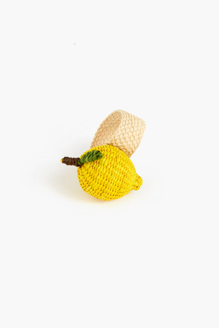 The Coro Cora woven napkin ring is handmade by women artisans in Colombia, featuring a yellow lemon with green leaf design. Crafted from natural fiber, it brings a charming touch to any table setting.