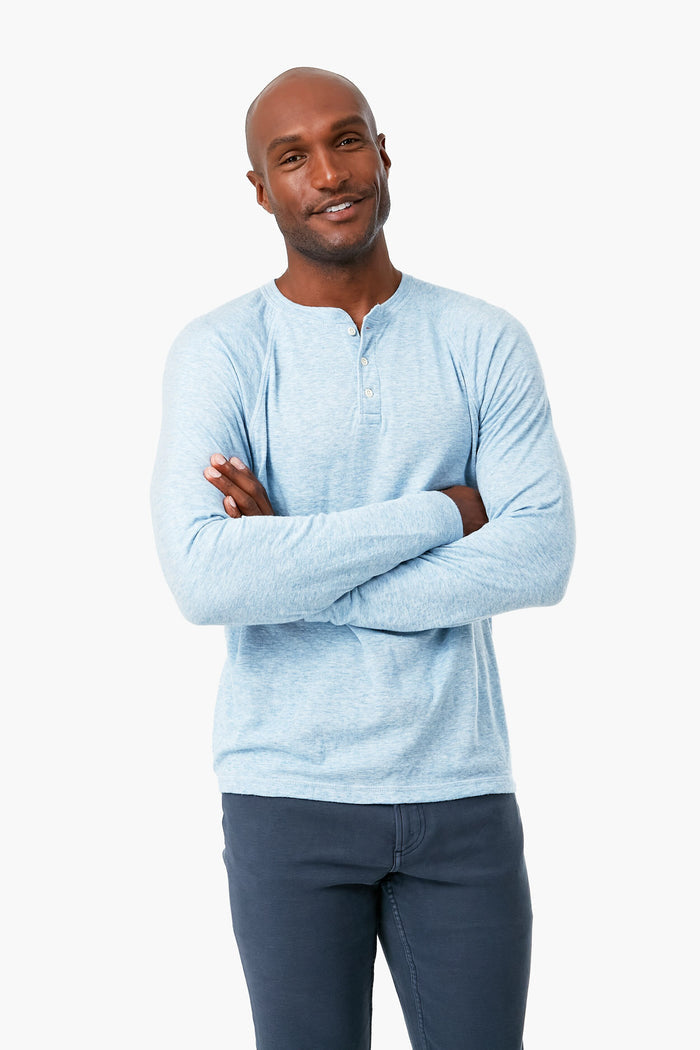 A man stands with arms crossed, wearing a light blue Faherty pima cotton long sleeve henley shirt and dark pants—a stylish and sustainable apparel choice.