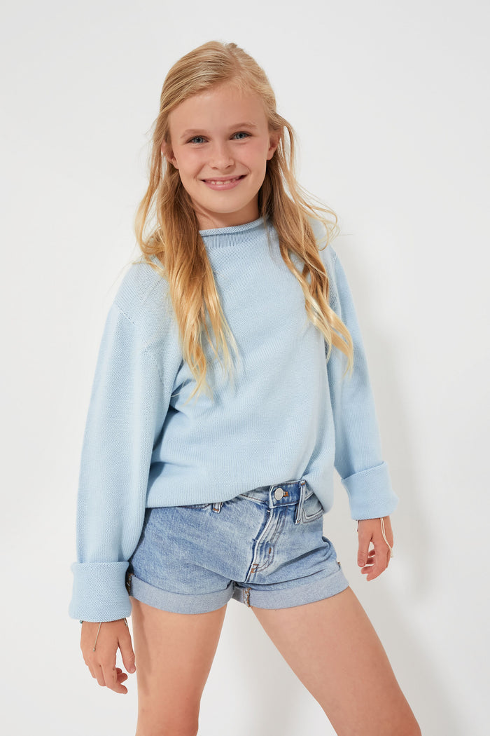 A girl smiles, wearing a light blue kids cotton sweater by A Soft Idea and denim shorts.