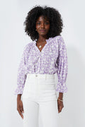 Woman wearing a lilac Ro's Garden shirt, a relaxed feminine blouse with ruffled cuffs, paired with high-waisted white pants.
