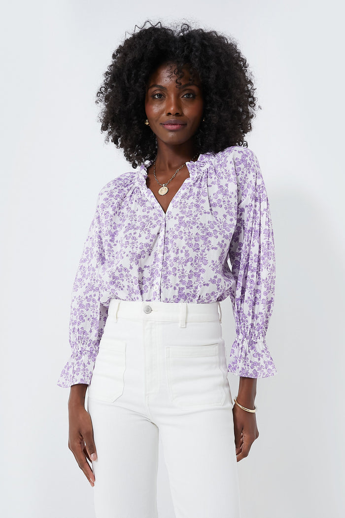 Woman wearing a lilac Ro's Garden shirt, a relaxed feminine blouse with ruffled cuffs, paired with high-waisted white pants.