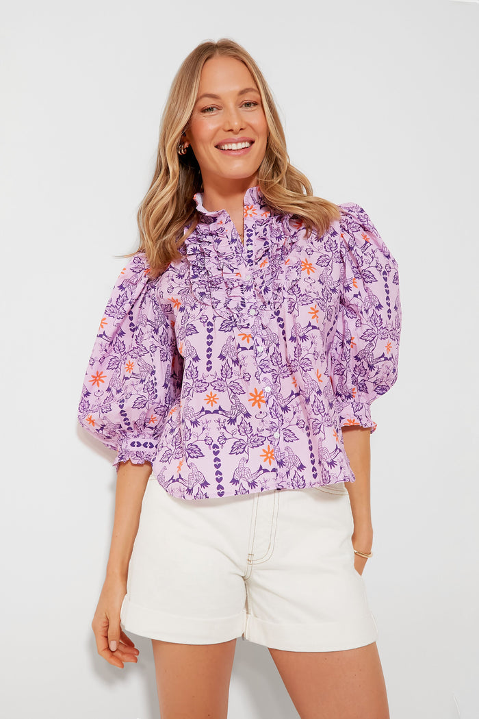 A woman smiles facing forward in an Oliphant Lovebird Lilac Blouse with a floral print and ruffled details, paired with white shorts.