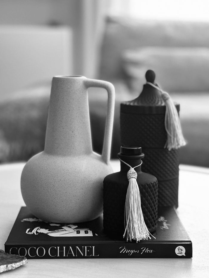 Black diffuser bottle beside a 3 stack complimenting candle with a grey coloured vase all on top of a black book.