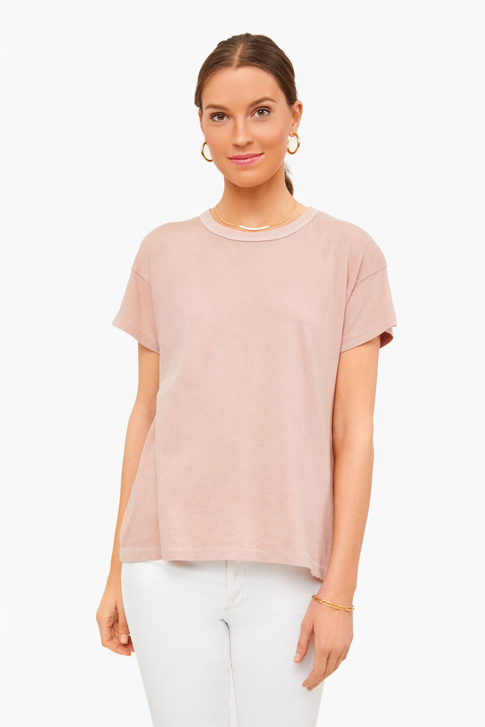 A woman wears THE GREAT.'s light pink boxy crew tee—a timeless staple—paired with white pants, hoop earrings, a necklace, and a bracelet.