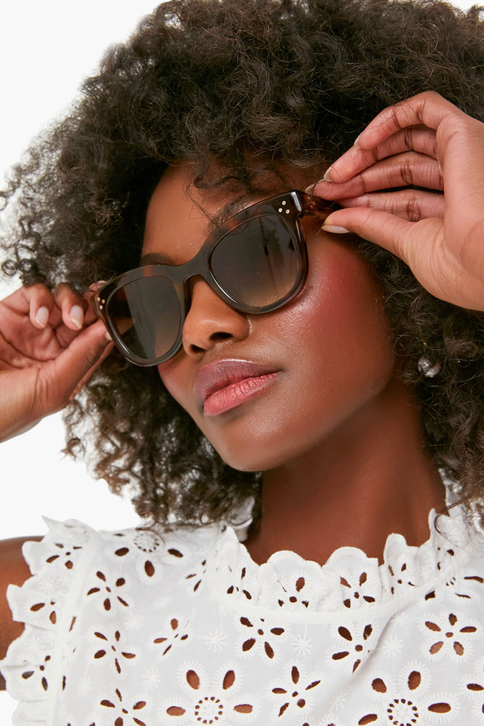 A woman with curly hair adjusts KREWE du optic dark sunglasses with UVA/UVB protection while wearing a white eyelet lace top.