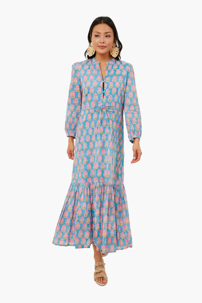 Woman wears a Love Tomas by Emerson Fry blue cotton dress featuring pink and yellow florals, long sleeves, a v-neck, and ruffled hem, styled with beige sandals and statement earrings.