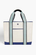 The Paravel tote is a beige canvas bag crafted from recycled materials, featuring blue accents, wide blue/black/white striped handles, a front pocket, and a snap closure.