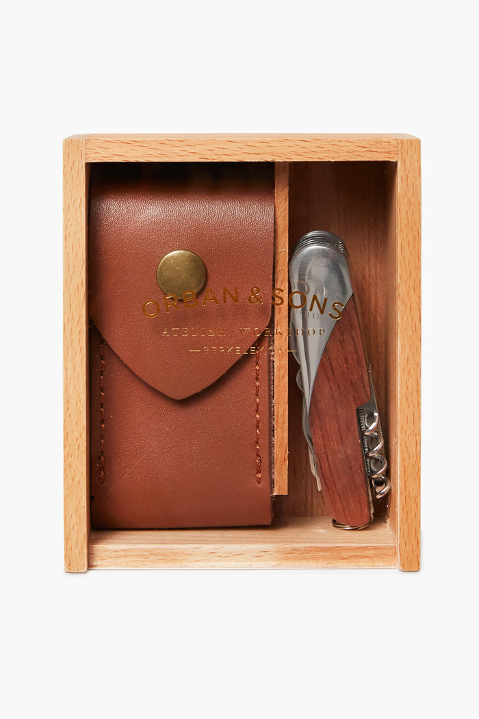 An Orban and Sons wooden box holds a brown leather carrying case with a brass button and a multi-function knife featuring a wooden handle.
