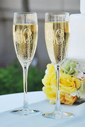 Two Carved Solutions personalized flute sets, each engraved with a monogram and filled with sparkling wine, sit on a table next to yellow and white flowers—an elegant gift for any celebration.