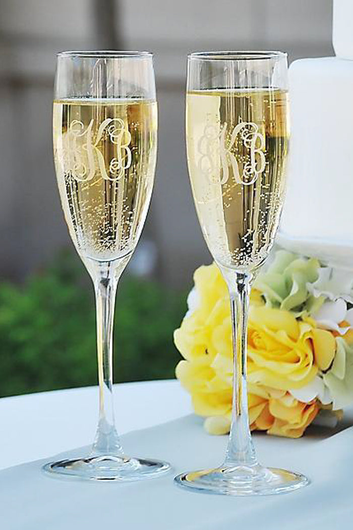 Two Carved Solutions personalized flute sets, each engraved with a monogram and filled with sparkling wine, sit on a table next to yellow and white flowers—an elegant gift for any celebration.
