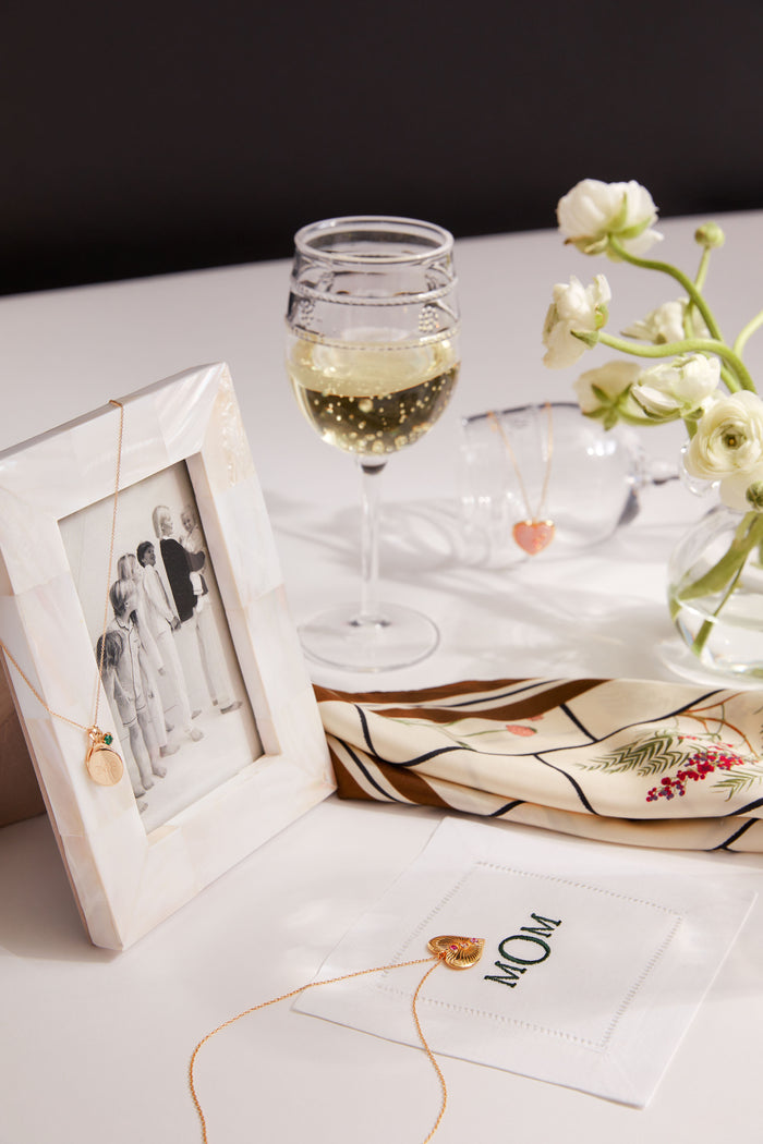 A Twos Company picture frame with a black-and-white family photo, jewelry, white wine, flowers, a scarf, and a MOM cloth are arranged on a white tabletop—perfect for a thoughtful housewarming gift.