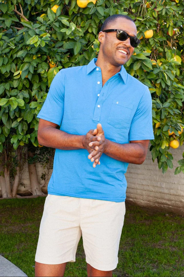 A man wearing a Criquet shirt and beige shorts stands outside by a citrus tree, smiling with his hands clasped.
