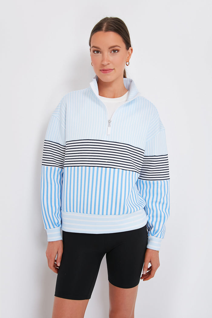 A woman wears an Addison Bay light blue and white Varsity Quarter Zip pullover with colorblocked stripes and black cycling shorts, showcasing stylish activewear.
