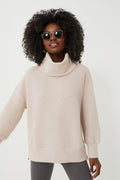 A woman with curly hair, wearing round sunglasses, a light beige ribbed turtleneck sweater from Varley, and dark pants, stands confidently in modern women's athleisure.