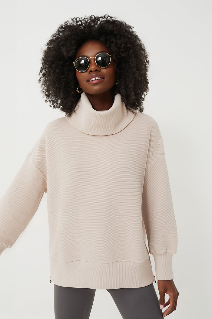 A woman with curly hair, wearing round sunglasses, a light beige ribbed turtleneck sweater from Varley, and dark pants, stands confidently in modern women's athleisure.