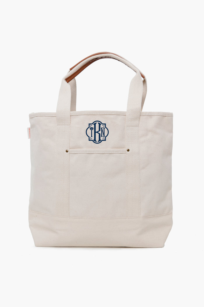 This CB Station cream canvas tote features short handles and a navy blue monogram on the front pocket, making it a stylish and eco-friendly reusable bag.