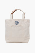 This CB Station cream canvas tote features short handles and a navy blue monogram on the front pocket, making it a stylish and eco-friendly reusable bag.