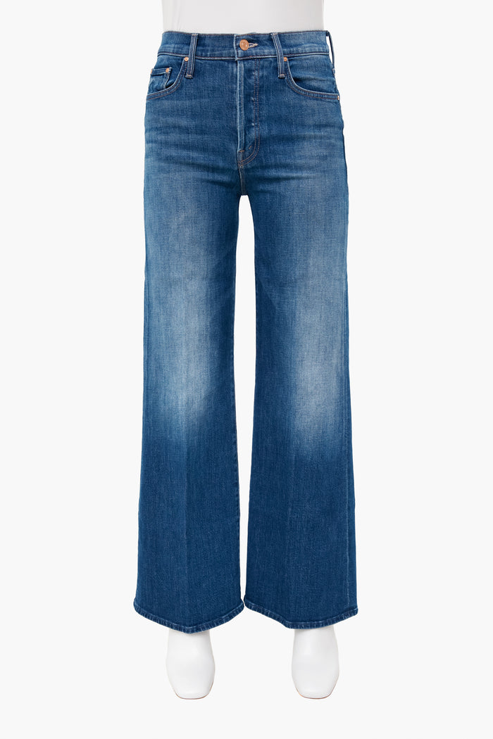 A pair of high-rise, wide-leg blue jeans with a faded wash by MOTHER, shown on a mannequin wearing white shoes.