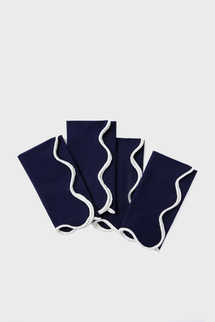 Four neatly folded Tuckernuck Home dark blue scalloped napkins with white wavy trim are arranged in a fanned-out pattern.