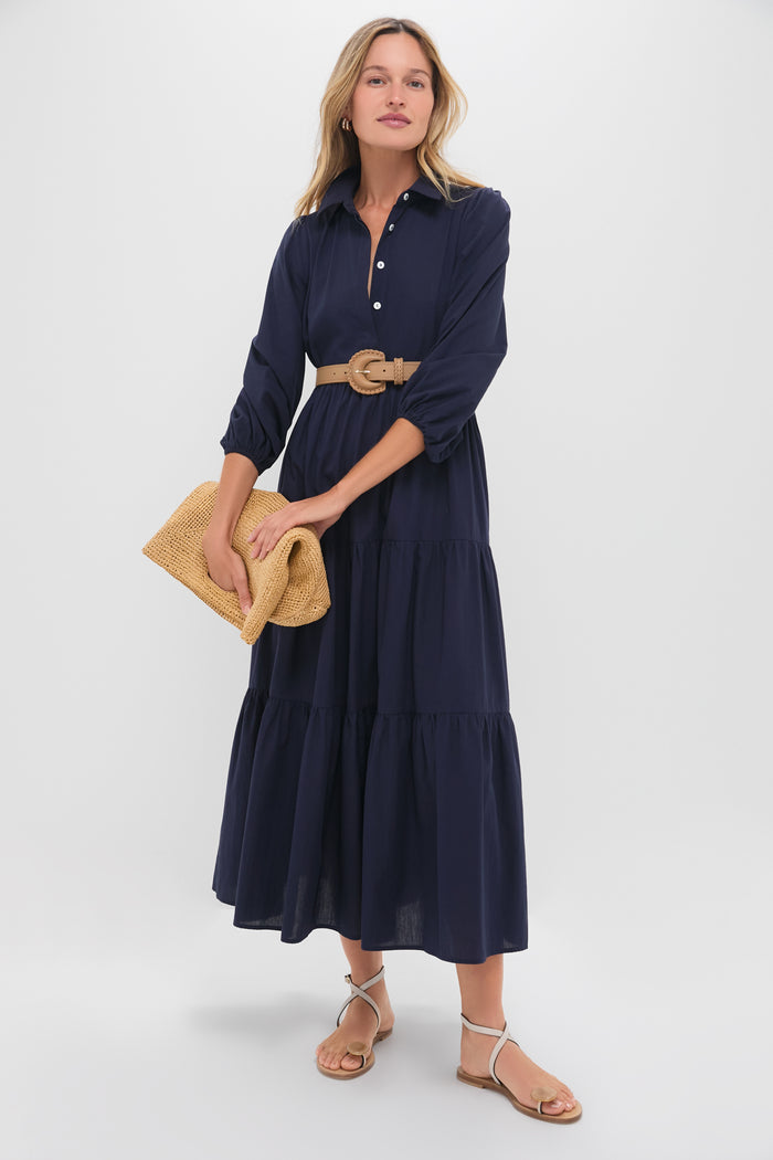A woman wearing a Pomander Place navy blue puff sleeve button-front maxi dress with a tan belt, holding a woven clutch, and wearing flat sandals.