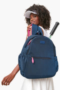 A woman in a white visor and dress carries the Ame & Lulu Courtside Backpack, crafted from lightweight navy nylon with pink zippers, featuring a tennis racquet in the side pocket along with other accessories.