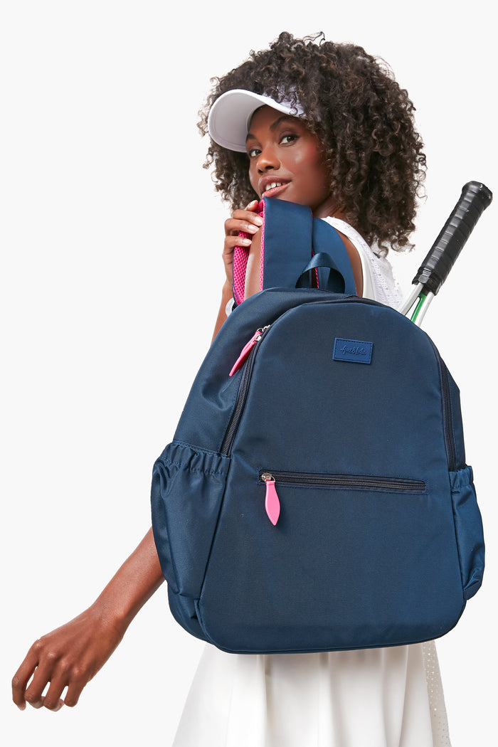 A woman in a white visor and dress carries the Ame & Lulu Courtside Backpack, crafted from lightweight navy nylon with pink zippers, featuring a tennis racquet in the side pocket along with other accessories.