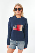 Smiling person wearing sunglasses, denim shorts, and a Tuckernuck Cropped Americana Sweater featuring an American flag pattern.