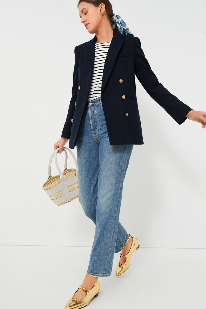 A person wears a Tuckernuck Navy Diana Blazer with gold buttons, a striped top, blue jeans, gold flats, and carries a woven handbag.