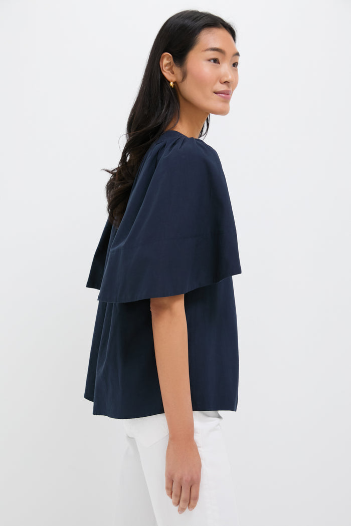 A woman stands sideways against a white background, wearing a Pomander Place loose-fitting navy blue flutter sleeve V-neck top and white pants.