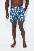 A man stands barefoot, wearing Fair Harbor blue men’s swim shorts featuring a white floral pattern and drawstring waist, made from upcycled plastic bottles.