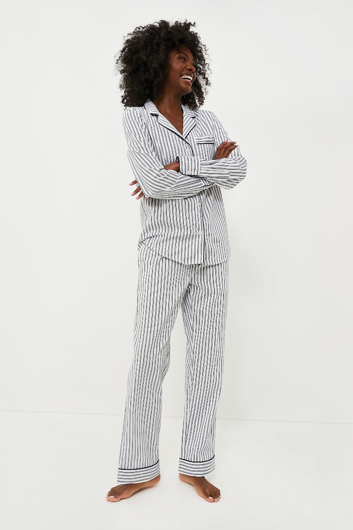 A woman stands barefoot, smiling with crossed arms, in a classic striped navy pajama set by Petite Plume. The long-sleeve, button-up cotton sleepwear offers a cozy and stylish look.